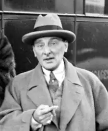 Middle aged, clean-shaven white man in hat and coat, smoking a cigar