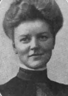 A young white woman with light hair piled on top of her head in a bouffant updo; she is wearing a high-collared dark dress or top