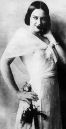 A young woman with light skin and dark hair parted center, standing, wearing a white dress with a translucent shawl; one hand is on her hip, and one hand is touching her hair or neck