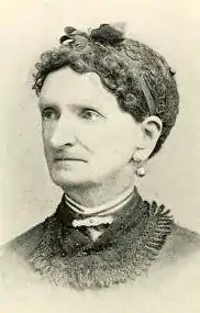 An older white woman, her hair caught in a snood with a bow on top; she is wearing a dark collar and drop earrings.