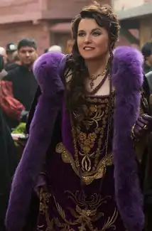 A pale woman with long brown hair wear an ornate purple outfit accented by golden stitching and a purple fur stole.