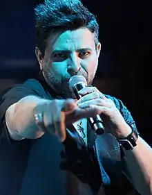 A young man with short black hair, singing with open arms.