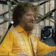 Picture of a man with goatee and curly hair, wearing a yellow shirt with white floral patterns. He is holding a guitar. Only the edges of the guitar can be seen in the photo. He is smiling away from the camera. It is a side profile photo.