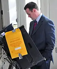 A young man with closely cropped hair and wearing a suit stands at an electronic voting machine