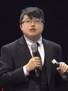 A person in a suit holding a microphone in their left hand with a red chair to their left.