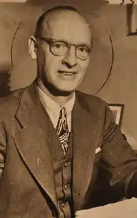 Lyle Wright in a suit and tie sitting a desk with a stack of papers.