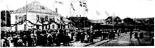 A black and white image showing a two-story building with a tiled hipped roof at left. It is surrounded by a crowd of people; where it thins out to the right of the image railroad tracks are visible. Above them fly flags with a bright cross on a dark background