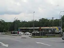 A small parking lot with many streetlights seen diagonally from across an intersection, with traffic signs in front of it, one of which is a red circle containing the number "50" on a yellow background. Behind the parking lot is a short modern-style gray, yellow and blue streamlined train underneath overhead wires.