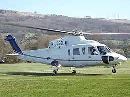 A white and blue helicopter on a grassy helipad, engines running and nose wheel lifted