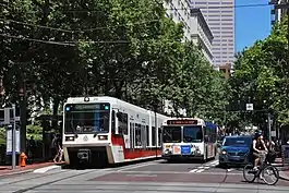 Photograph of a Max train on the left, a bus in the center and a woman on a bicycle making a turn off the Portland Transit Mall