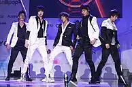 Five men with bowl haircuts and eyeliner wearing close-fitting, shiny suits—some black with white embellishment, others white with black embellishment.