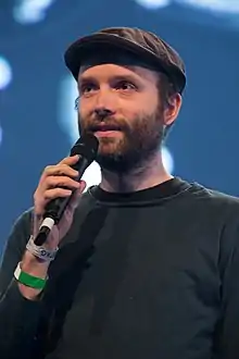 bearded White male wearing a flat cap and a dark long-sleeved shirt, holding up a microphone and looking right of camera