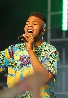 A black man wears a colourful shirt and sings into a microphone