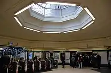 A station ticket hall with automatic ticket gates on the left and passengers waiting to buy tickets from a machine in the wall. A large octagonal roof light occupies the central portion of the ceiling with a deep convex moulding around the opening.