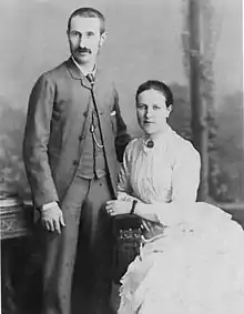 photo of man dressed in suit standing with woman in white dress seated beside him