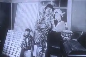 A young girl, man, and woman standing outside of a house, all looking up in the sky. The girl, on the left, is smiling and pointing skyward. The man wears a bowler hat and holds a short broom over his shoulder; the woman wears a kerchief around her head. They are surrounded by domestic objects as if just moving into or out of the house.