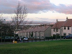 A row of housing on a downhill street with bare trees lining the opposite side, a view of the Forth Valley and Ochil Hills in the background.