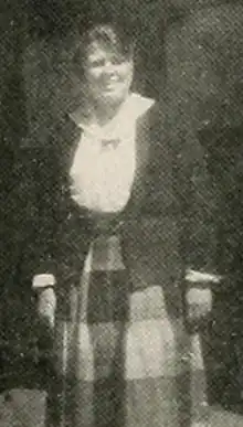 A young white woman, standing and smiling, wearing a white blouse, a dark cardigan or jacket, and a full skirt with a graphic pattern