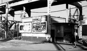 A monochrome image of an apparently-white brick building from a three-quarter view from the building's proper right. The building has a semicircular bay in its front. Behind the building is an arched steel lattice