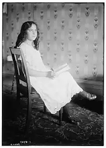 A white girl with loose dark hair, seated sideway on a wooden chair; she is holding a book and wearing a loose-fitting white dress.