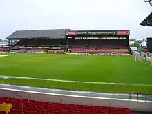 In the foreground, a grass football field. In the background, a two-part seating area. Both parts are covered and are connected, but the roofs are distinctly different.