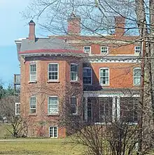 The same house seen from the side with the large wing, also in springtime, illuminated by sun from the left, with some bare branches and bushes partially obscuring the view
