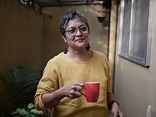 Older South Asian woman with short hair wearing a yellow sweater and glasses, holding a red mug in one hand, smiling at camera