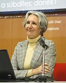 Portrait photograph of an old woman with short gray hair, wearing a beige turtleneck and a plaid suit jacket, arms crossed, standing in front of a microphone and podium.
