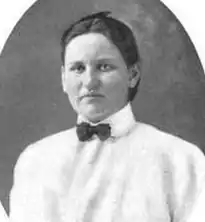 White woman, hair smoothed close to head and back behind neck, wearing a white blouse and a small bowtie.