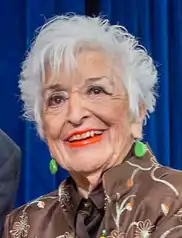 An older white woman with spiky short white hair, wearing bright green drop earrings and an embroidered jacket