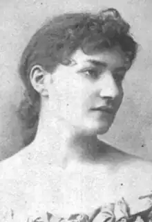 A white woman with dark hair dressed to the nape, with a curly fringe; she is wearing a low-cut gown with bare shoulders