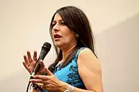 A brunette Caucasian woman wearing a blue blouse is holding a microphone.