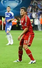 Gómez, in a Bayern kit, stands on the pitch looking upwards with a Chelsea player and fans in the background