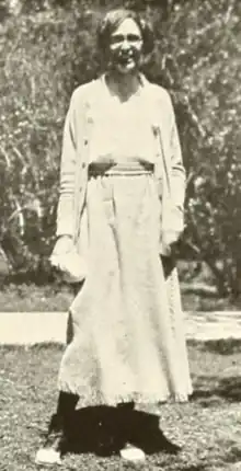 A young white woman, standing outdoors and smiling, wearing a light-colored loose-fitting skirt, jacket, and blouse