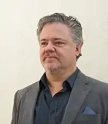 Head and shoulders photographic portrait of a white man with greying hair and a short beard and moustache in an open-necked dark blue shirt and grey jacket