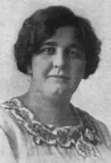 A black-and-white photograph of a white woman with dark wavy hair and a dimpled chin; she is wearing a dress or blouse with an embroidered square neckline