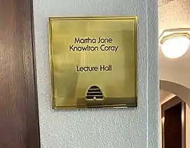 a gold plaque affixed to a wall that reads "Martha Jane Knowlton Coray Lecture Hall." Under the inscription is a graphic of a beehive in black.