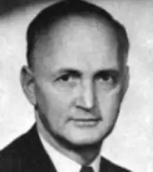 A middle-aged balding white man; he is cleanshaven and wearing a coat and tie