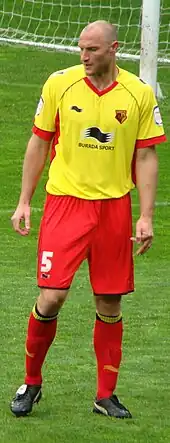 A man wearing a yellow shirt, red shorts and red socks, standing on a grass field. A goalpost and net are visible behind him.