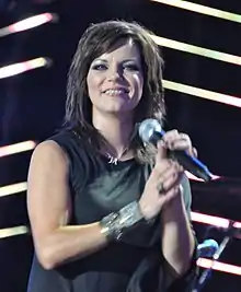 A young woman with long dark hair wearing a dark sleeveless t-shirt, holding a microphone