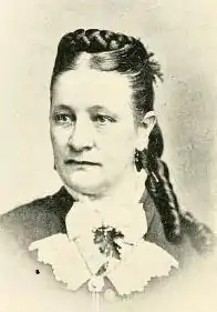 A white woman with her dark hair braided across the crown, and long curls behind her ear.