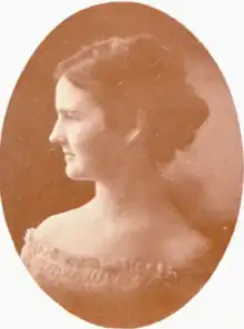 A young white woman with dark hair, in profile, wearing a light-colored dress with an off-the-shoulders smocked neckline; in an oval frame