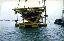 A partial section of a wooden ship lying inside a massive steel frame is suspended just about the water with a partial view of a barge on the left and small boats in the background.