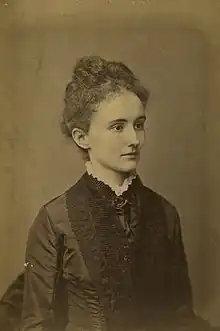 A young white woman with hair arranged in an updo, wearing a heavily embroidered dark dress with a high lace-edged collar