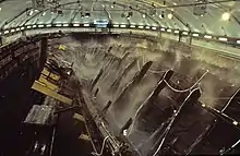 A diagonal section of a wooden ship is lying on its side steel frame at an angle, being sprayed with water from dozens of sprinklers