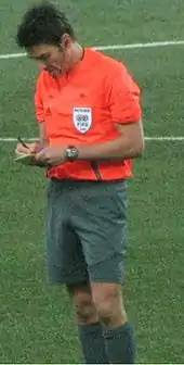 A man wearing a bright orange shirt and grey shorts, writing in a notebook. He has dark hair.