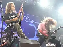 A color photograph of two members of the group Children of Bodom standing on a stage with guitars, drums are visible in the background. Both electric guitarists have "flying V" style guitars and they have long hair.