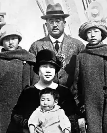 A Japanese family of five, posed outdoors together: the father is standing, wearing and glasses; the two daughters are standing, wearing hats and coats; the mother is seated, wearing a dark hat, holding a baby boy