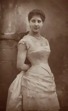 Young white woman with fairly short, dark hair, in white or cream Victorian frock, leaning against a pillar