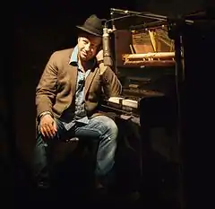 Mauro Scocco wearing faded blue jeans, a light blue shirt and brown jacket, sitting sideways next to a piano, head propped up on one elbow, smiling and looking right of camera